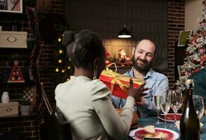 Diverse friends offering gifts to each other for christmas holiday, spreading positivity at festive dinner. Happy man giving present box to african american woman during winter celebration. photo