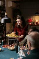 Woman serving festive meal at table, celebrating christmas dinner with diverse friends and family at home. People feeling joyful eating homemade food and drinking wine during xmas eve event. photo