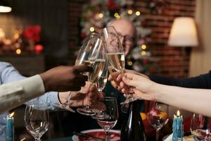 Happy persons clinking glasses at christmas eve festivity, celebrating winter holiday with alcohol and homemade food. Diverse friends and family saying cheers at table, cozy decor. photo
