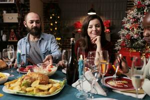 joven Pareja celebrando Navidad con familia, mujer asistiendo festivo cena durante Navidad víspera fiesta a hogar. personas disfrutando tradicional comida en el mesa, sensación alegre y alegre. foto