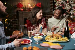 Diverse people having fun at dinner on christmas eve, celebrating december holiday around the fireplace at home. Friends enjoying celebration surrounded by festive decor and xmas tree, feeling jolly. photo