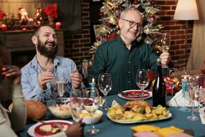 mayor hombre haciendo brindis con elevado vaso de vino a Navidad víspera cena a celebrar diciembre fiesta con amigos y familia. abuelo haciendo habla alrededor el mesa, espumoso alcohol. foto