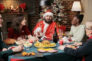 Santa claus cosplay spreading positivity around friends, gathering at dinner table to celebrate christmas eve event. Young adult dressed in santa suit feeling jolly during seasonal december festivity. photo