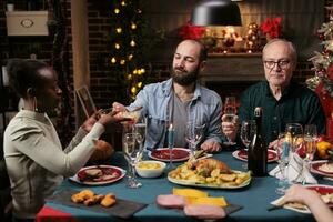 Diverse friends gathering at home to celebrate seasonal winter holiday, feeling jolly during christmas eve dinner festivity. People and family enjoying food and wine glasses, december tradition. photo