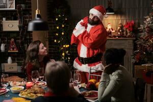 Santa claus man carries red sack filled with gifts and presents, ensuring amazing christmas eve festive dinner with friends and family at home. Group of people feeling joyful with man in santa hat. photo