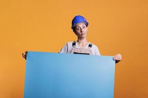 Industrial builder holding blank cardboard sign for advertisement in studio. Optimistic renovating contractor in uniform showing empty carton banner with isolated copyspace, mockup. photo