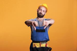 Displeased handyman in overalls showing thumbs down, expressing dislike and disapproval symbol over studio background. Male constructor doing disagreement gesture, being disappointed. photo