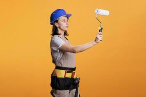 Young constructor holding paintbrush roller and painting walls in studio, posing with brush. Female painter working on painting and renovating job, using tool for redecoration improvement. photo