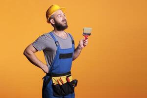 Young painter decorator painting walls with brush in studio, posing with paintbrush and tools before working on renovation. Craftsman expert using brush and color to do diy project. photo