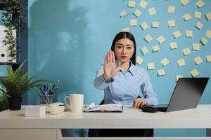 Woman seated at office desk showing rejection with the palm of her hand, expressing dissatisfaction with working conditions. Employee refusing to receive more workload without rest. photo