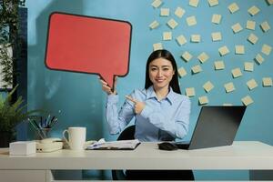 Professional asian woman with advertising speech bubble board against blue wall, showing isolated cardboard text message. Corporate employee holding poster with copy space. photo