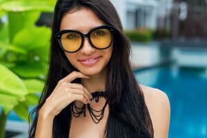 Sensual woman with perfect slim tan body in trendy necklace , black swimwear and transparent beach cover up , trendy yellow sunglasses  enjoying  holidays  near amazing pool in tropical resort. photo