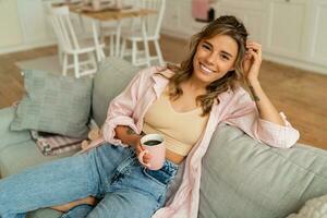 Lovely woman in casual clothes resting un sofa in cozy  living room. Holding cup of  cappuccino. photo