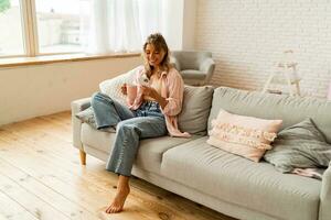 Happy woman drinking coffee in early morning, sitting on sofa in pink  shirt at home. Using smartphone. photo