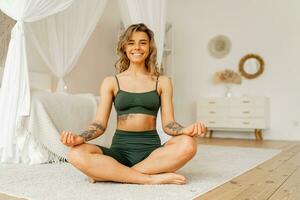 hora para yoga. bonito joven mujer hacer ejercicio y sentado en yoga loto posición mientras descansando a hogar. elegante escandinavo interior. foto