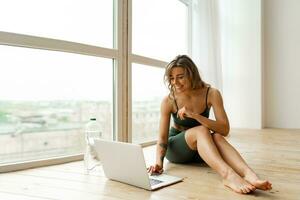 Pretty  woman in sportswear is sitting on the floor with  bottle of water and is using a laptop at home in the living room. Sport and recreation concept. photo