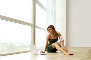 Pretty  woman in sportswear is sitting on the floor with  bottle of water and is using a laptop  in  modern fitness  studio . Sport and recreation concept. photo