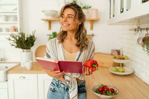 Funny housewife woman looking at recipe in cookery book preparing vegetable salad cooking food in light kitchen at home. Dieting healthy lifestyle concept. photo