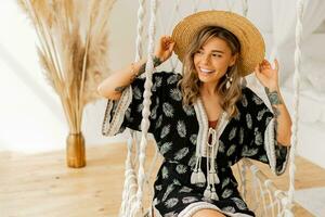 Cozy home atmosphere.  Lovely stylish blond woman in boho dress posing in stylish bedroom. Ladysitting on hanging swing . photo