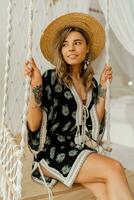 Smilling young woman in boho dress posing in stylish bedroom.  Sraw hat, boho feather drop earrings . Lady sitting on hanging swing. photo