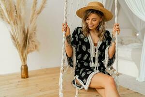 Smilling young woman in boho dress posing in stylish bedroom.  Sraw hat, boho feather drop earrings . Lady sitting on hanging swing. photo