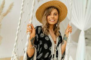 Smilling young woman in boho dress posing in stylish bedroom.  Sraw hat, boho feather drop earrings . Lady sitting on hanging swing. photo