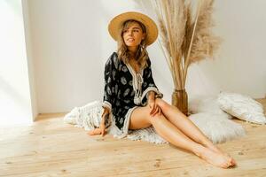 Stylish  blond woman in straw hat and boho dress  posing in studio over white background  with pampas grass decor.  Pillows with macrame ornament. Summer trends. photo