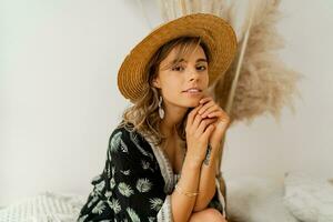 Close up portrait of woman in bohemian dress sitting on pillows at home. Wearing earrings with feather  and bracelets. White background in studio with pampas grass decor. photo