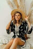 Lovely young woman in bohemian dress sitting on pillows at home. Wearing earrings with feather  and bracelets. White background in studio with pampas grass decor. photo