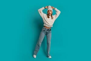 Studio winter portrait of  smiling blond woman in white hat and whool sweater  send kiss to camera ,posing in studio over turquose background. Cold season acsessories and fashion.  Full lenght. photo