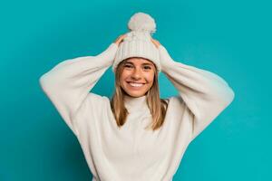 Studio winter portrait of  smiling blond woman in white hat and whool sweater posing in studio over turquose background. Cold season acsessories and fashion. photo