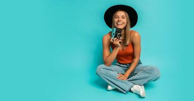 Lovely blond woman in  stylish summer outfit holding retro camera , sitting on floor in studio on blue background. Vacation mood. photo