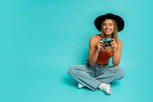 Lovely blond woman in  stylish summer outfit holding retro camera , sitting on floor in studio on blue background. Vacation mood. photo