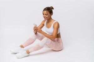 Fitness and lealthy lifestyle concept. Happy sporty young woman sitting on the floor at studio after exercising, and using smart phone. White background. photo