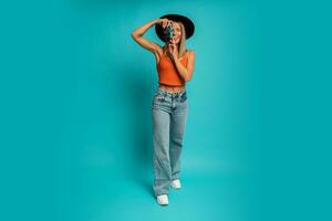 Pretty laughing woman in black hat and orange top  holding vintage camera and posing over turquoise background in studio.  Full lenght. photo