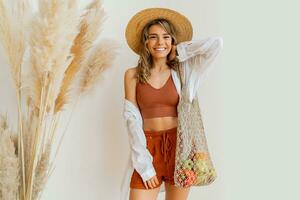 Smiling  blond woman with wavy hairs holding eco friendly mesh shopper with fresh  vegetables  in studio with boho interior. photo