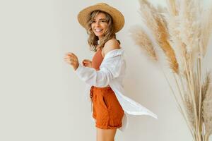 Boho mood. Stylish woman in summer outfit , straw hat  and orange clothes posing over white background in studio with pampas grass decor. photo