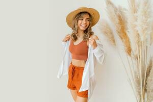 Boho mood. Stylish woman in summer outfit , straw hat  and orange clothes posing over white background in studio with pampas grass decor. photo