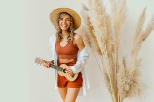 Indoor  photo of  blond graceful woman in  summer outfit with straw hat  playing ukulele guitare  over white background in studio with pampas grass decor.