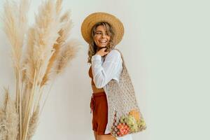 Pretty blond woman in straw hat  holding eco friendly mesh shopper with fresh  vegetables  in studio with boho interior. photo