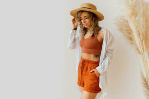 Boho mood. Stylish woman in summer outfit with straw hat posing over white background in studio with pampas grass decor. photo