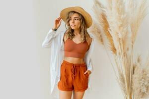 Boho mood. Stylish blond  woman in summer outfit with straw hat posing over white background in studio with pampas grass decor. photo