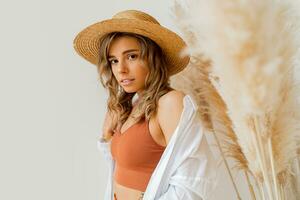 Close up portrait of attractive  woman in summer outfit with straw hat posing over white background in studio with pampas grass decor. photo