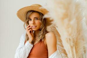 Close up portrait of attractive  woman in summer outfit with straw hat posing over white background in studio with pampas grass decor. photo