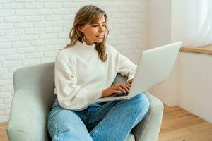 Pretty  woman with blond wavy hairs sitting on sofa at home working on laptop computer.   Wearing cozy sweater. photo