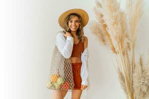 Smiling  blond woman with wavy hairs holding eco friendly mesh shopper with fresh  vegetables  in studio with boho interior. photo