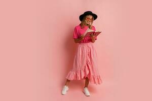 Cute blond woman in black hat and pink spring outfit holding book and  posing in studio.  Full lenght. photo