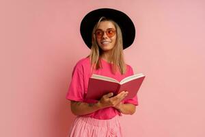 Cute blond woman in black hat and pink spring outfit holding book and  posing in studio. photo