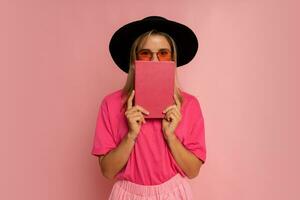 Cute blond woman in black hat and pink spring outfit holding book and  posing in studio. photo