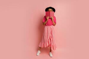 Cute blond woman in black hat and pink spring outfit holding book and  posing in studio.  Full lenght. photo